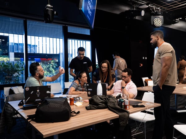 A group of people are gathered around a wooden table in a modern indoor workspace, engaged in a collaborative discussion with laptops and drinks present.