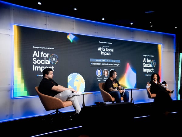 A panel discussion at a tech event titled 'AI for Social Impact', featuring three speakers on a stage in front of a large digital screen.