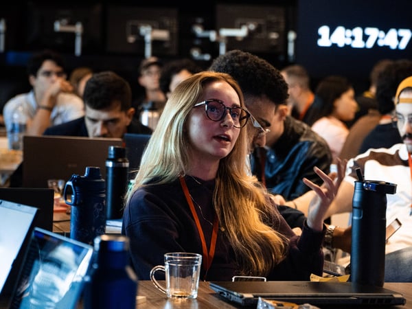 A young woman with blonde hair and glasses is captured mid-speech or gesturing during a workshop or collaborative event, surrounded by other participants and laptops.