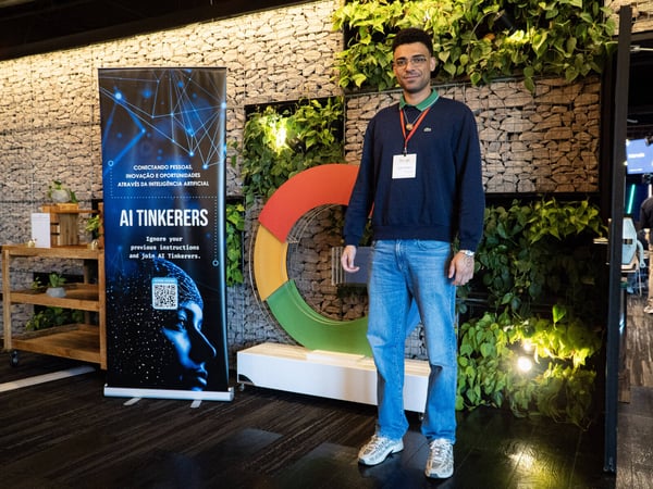 A man stands in an indoor event space next to an 'AI Tinkerers' banner and a large Google logo sculpture against a stone wall with plants.