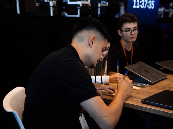 A group of young individuals are seated at a wooden table, focused on writing on sticky notes and working with laptops in a dimly lit indoor environment.