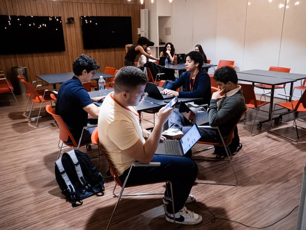 A group of young adults are working on laptops and mobile devices in a modern coworking space with orange chairs and wood-paneled walls.