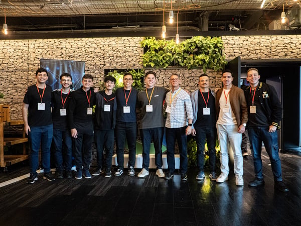 A group of ten men posing for a portrait in an indoor space featuring a textured stone wall and hanging light bulbs.