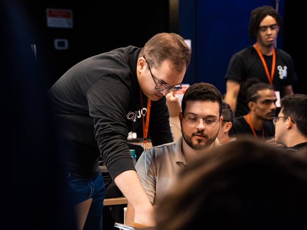 A man in a black hoodie leans over to assist another man seated at a desk in a dimly lit indoor environment, likely during a tech event or workshop.