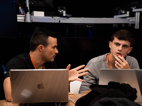 Two men are sitting at a wooden table with laptops, engaged in an active discussion where one is gesturing and the other is listening intently.
