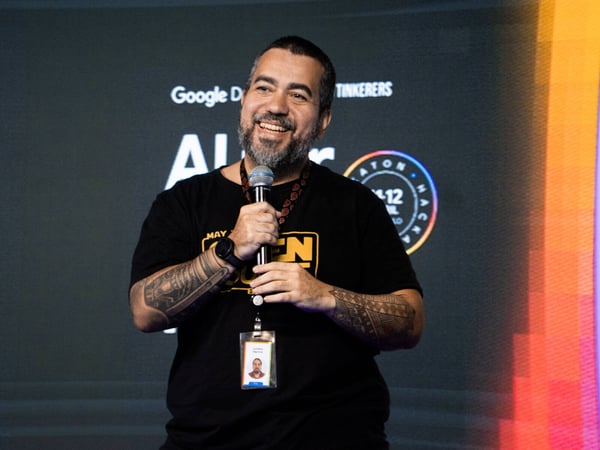 A man with a beard and tattoos is smiling while holding a microphone on a stage at a Google Developers event.
