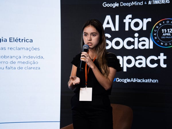 A young woman is speaking into a microphone while giving a presentation at an 'AI for Social Impact' hackathon event.