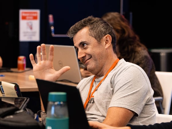 A man in a grey t-shirt and orange lanyard smiles and holds up his hand while sitting at a desk with laptops in a workspace setting.