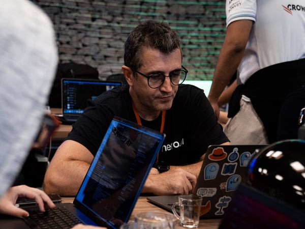 A man with glasses and a black t-shirt is intently focused on a laptop screen in a busy indoor environment filled with other tech equipment and people.