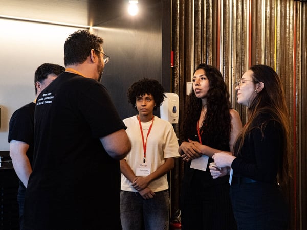 A group of five people are gathered in an indoor space, engaged in a conversation. A man with a message on his shirt stands with his back to the camera, facing three women and another man partially visible on the left.