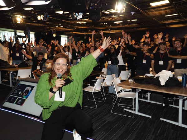 A woman in a green shirt smiles and holds a microphone in front of a large crowd of people with their hands raised in an indoor event space.