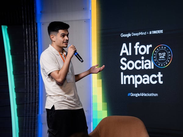 A man stands on a stage holding a microphone, giving a presentation in front of a screen that reads 'AI for Social Impact'.