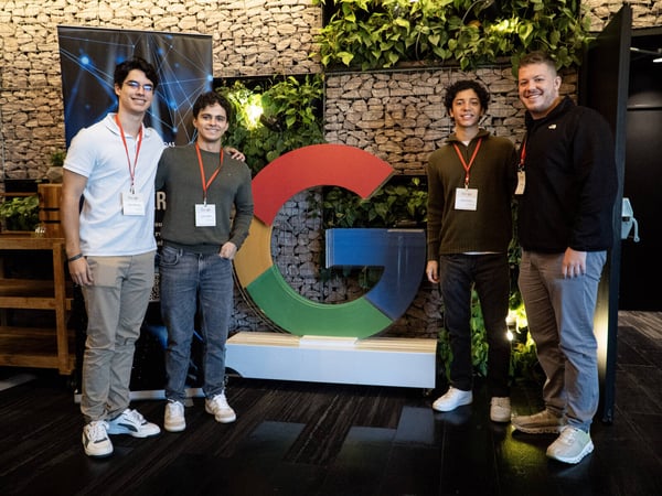 Four young men are posing for a group photo in front of a large, colorful Google 'G' logo in an indoor office setting.