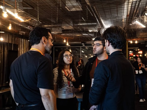 A group of four people are engaged in a conversation in an indoor space with an industrial ceiling and warm, hanging lights.