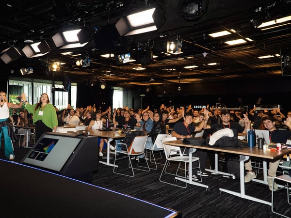A group of people attending a workshop or presentation in a dark studio-like environment equipped with professional lighting. Two women at the front are high-fiving while the seated audience members raise their hands in participation.