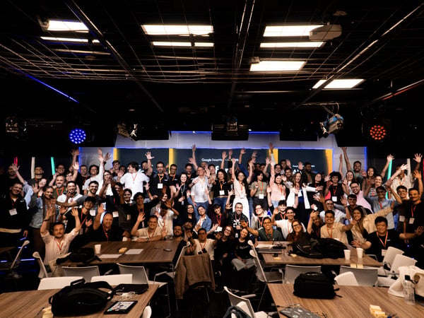 A large group of people posing for a group photo in an indoor event space, with many individuals waving towards the camera.
