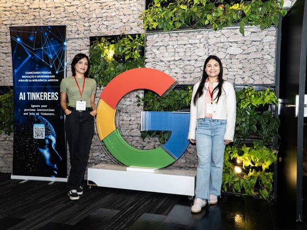Two women pose for a photo in an indoor space featuring a large Google 'G' logo, a stone wall with greenery, and an 'AI Tinkerers' banner.