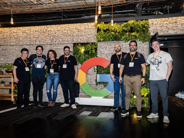 A group of seven people posing for a commemorative photo in front of a large, colorful Google 'G' logo and a stone gabion wall decorated with plants.