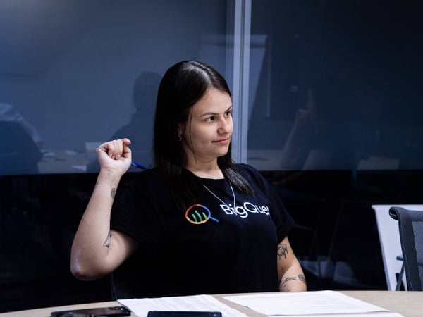 A woman wearing a black BigQuery t-shirt is sitting at a table in an office setting, looking off-camera while holding a pen.