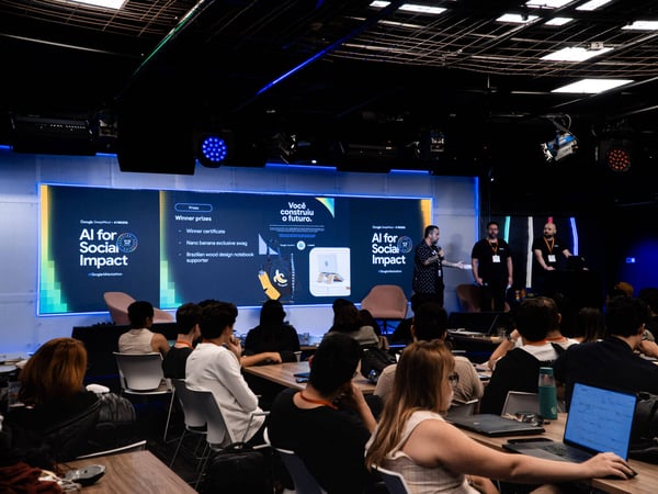 A photograph of a presentation event titled 'AI for Social Impact' by Google DeepMind and STRIDE, featuring speakers on a stage and an audience seated at tables.