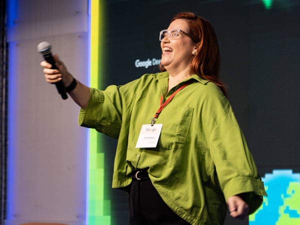 A woman with red hair and glasses is laughing joyfully while holding a microphone on a stage during a presentation.