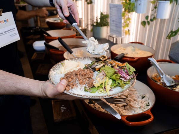 A close-up photograph of a person serving themselves food from a buffet line, adding a creamy dish to a plate already containing rice, beans, and salad.