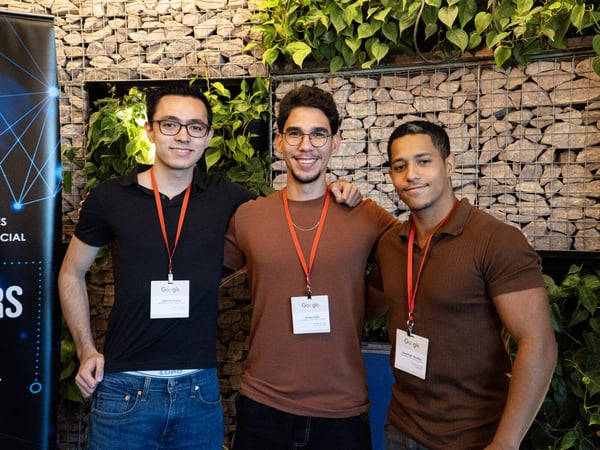Three young men wearing Google lanyards stand together for a photo in front of a stone wall decorated with green plants.