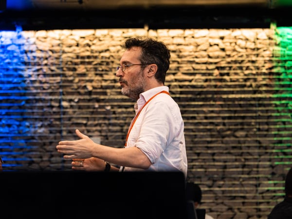A man with a beard and glasses is seen in profile, gesturing with his hands while speaking in front of a textured stone wall with colored lighting.