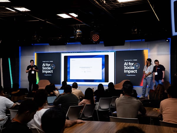 A presentation or demo session at an 'AI for Social Impact' event, featuring a speaker on stage and an audience with laptops.