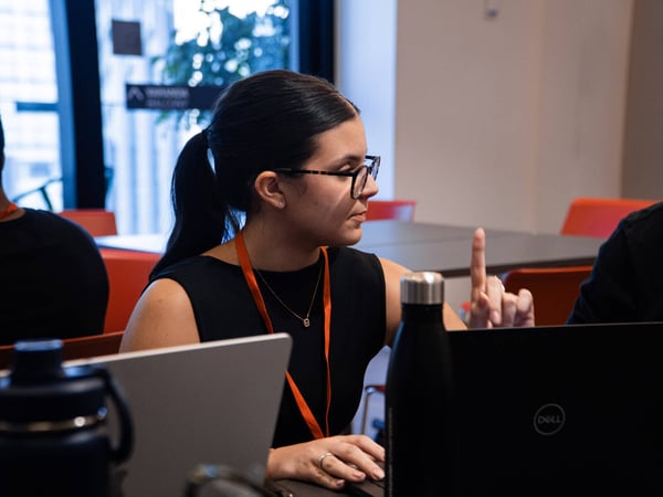 A woman with glasses and a ponytail is captured in a candid moment, gesturing with one finger while sitting at a table with laptops.