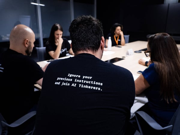 A group of people are seated around a conference table during a meeting, with the person in the foreground wearing a t-shirt with text about AI.