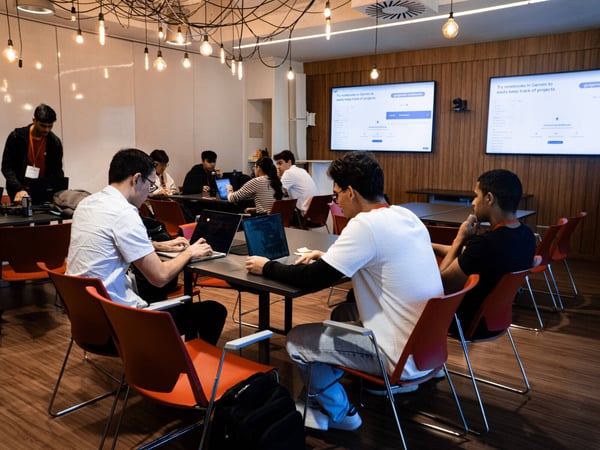 A group of people are working on laptops at tables in a modern, well-lit indoor space with large digital screens displaying project management information.