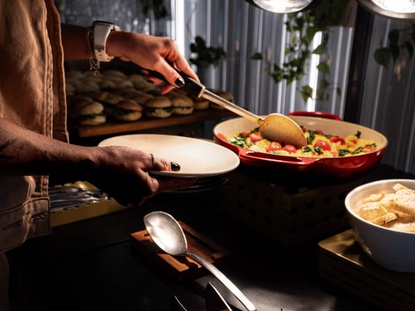 A close-up photo of a person serving food from a large red skillet onto a plate at a buffet-style event.