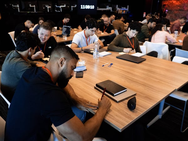 A group of people are seated at long wooden tables in a dimly lit room, focused on writing in notebooks or working on laptops, with a digital clock visible in the background.