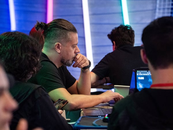 A man with a man-bun and beard is seen in profile, deeply focused on a laptop screen in a dimly lit room with vibrant neon tube lights.