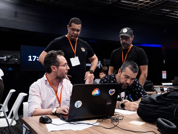 A group of men are collaborating around a laptop at a wooden table, with some standing and others seated in what appears to be a workshop or tech event.
