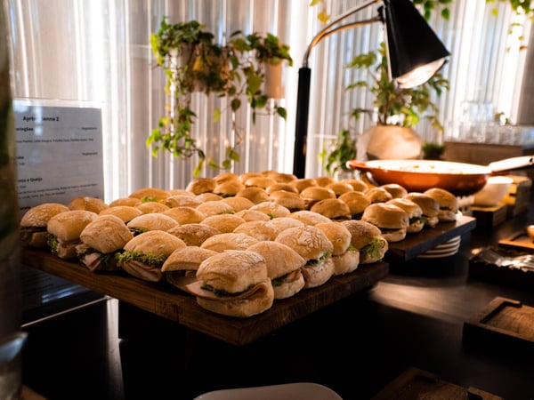 A catering buffet display featuring rows of small sandwiches on wooden boards, illuminated by a warm heat lamp in an indoor setting.