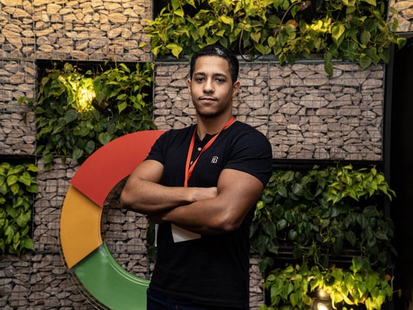 A young man stands with his arms crossed in front of a decorative wall made of gabion stones and lush green plants, next to a large, colorful logo element.
