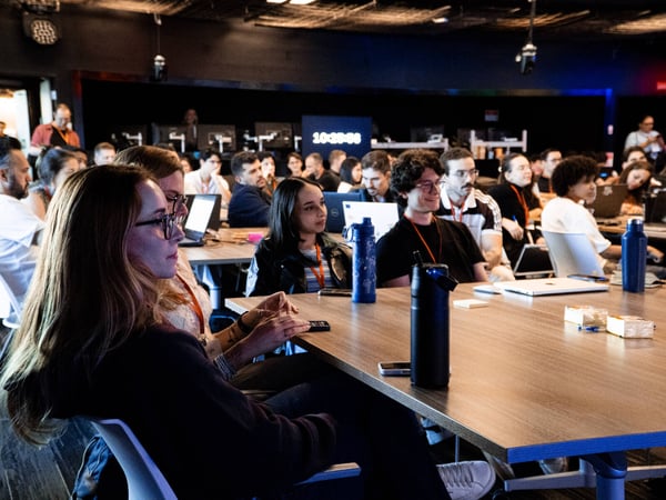 A group of people sitting at long tables in a dimly lit conference or workshop setting, looking toward a presentation area.