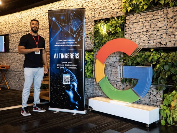 A man stands smiling next to an 'AI Tinkerers' banner and a large, colorful Google 'G' logo in an indoor space with a stone wall and green plants.