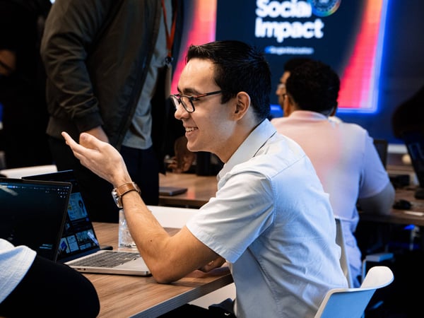 A man in a light blue shirt and glasses is captured in a candid moment, gesturing during a discussion at what appears to be a tech event or office setting.