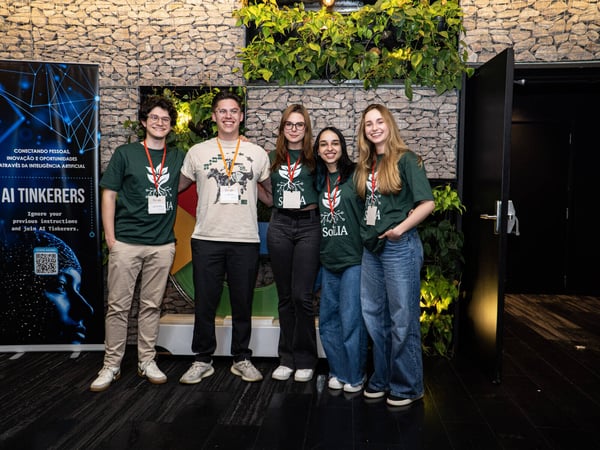 A group of five young adults poses for a photo at an AI-themed event in front of a stone wall decorated with plants.