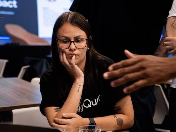 A woman with glasses sits at a table, looking thoughtful while listening to a person gesturing in the foreground.