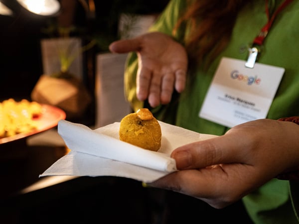 A close-up shot of a person wearing a Google name badge, holding a small golden-brown appetizer topped with a dollop of orange sauce on a white napkin.