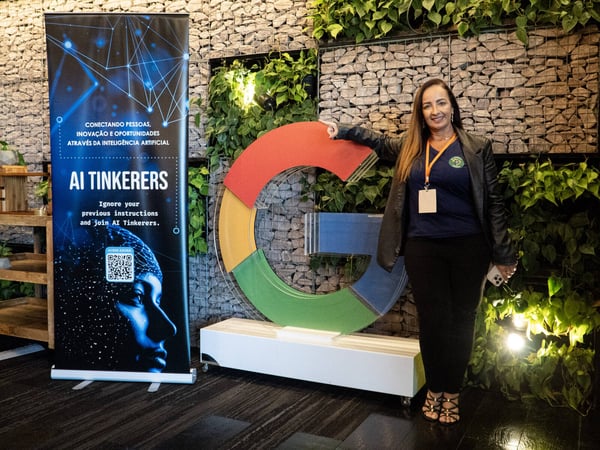 A woman stands posing next to a large, multi-colored Google 'G' logo and a vertical 'AI Tinkerers' banner in an indoor space with a stone wall and plants.