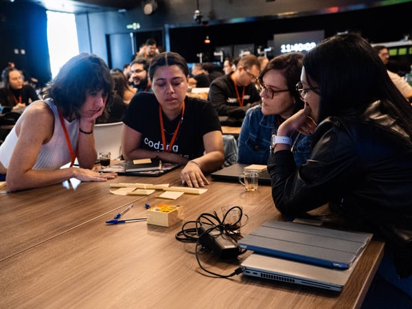 A group of women are gathered around a wooden table in a large room, focused on a collaborative activity involving sticky notes and pens.