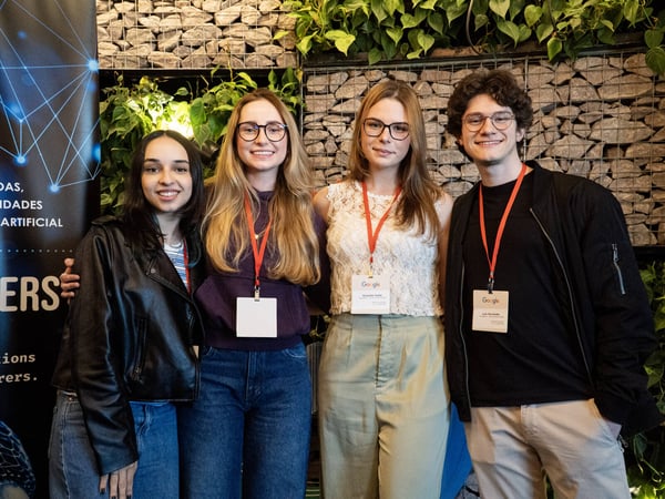 A group of four young adults, three women and one man, posing for a photo at an event in front of a stone and plant wall.