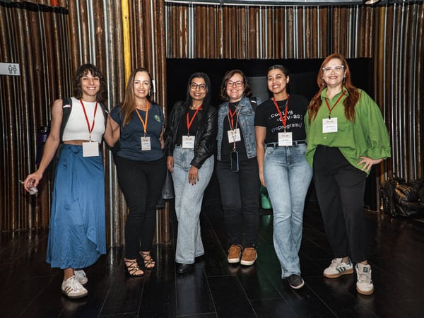 A group of six women smiling and posing for a photo indoors at an event, all wearing lanyards with identification badges.