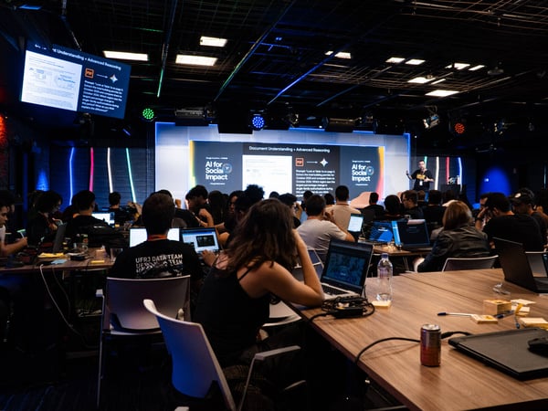 A speaker presents on a stage about AI for Social Impact to a large audience of people seated at tables with laptops in a modern, dimly lit event space.