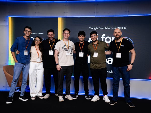 A group of seven people pose for a photograph in front of a large digital screen at an event hosted by Google DeepMind and AI Tinkerers.